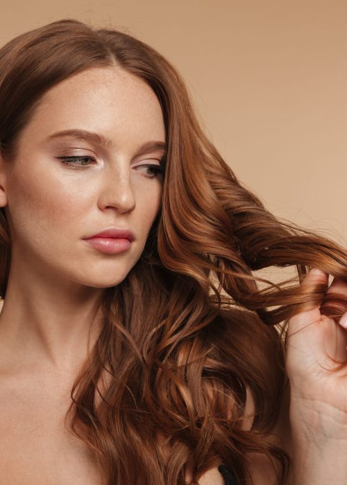 Beauty portrait of pretty ginger woman with long hair touching her hair and looking away over cream background
